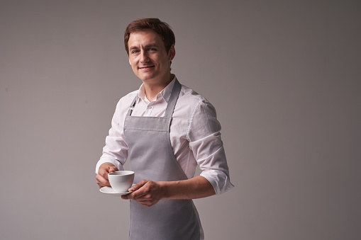 The positive male waiter on a grey background. The barista holds a coffee drink for the customer. Portrait of a successful young man. Layout with copy space. High quality photo