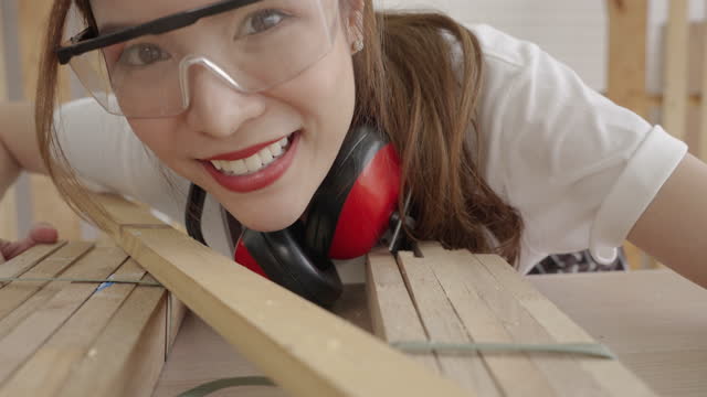 Beautiful Asian female carpenter blowing wood chips with air from her mouth out of planks.