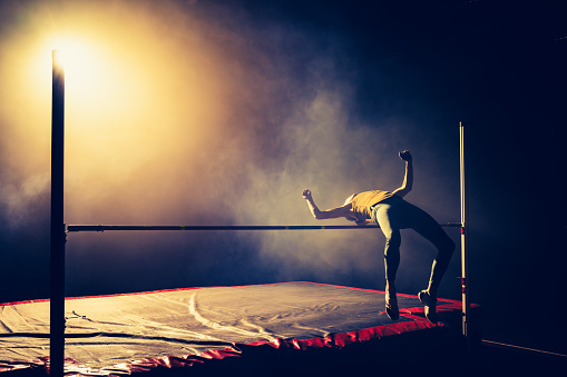 Sportsman silhouette training high jump and flying through pole illuminated in dark gym wide shot