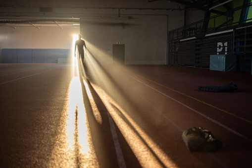 Sportsman walking at dark indoor stadium with sunlight shining through door back view wide shot