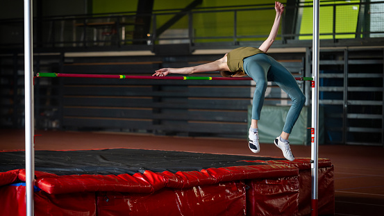 Sportswoman high jumping through pole and falling on exercise mat while training in gym wide shot