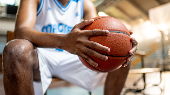 Male basketball player holding ball in hands and sitting on bench at indoors court