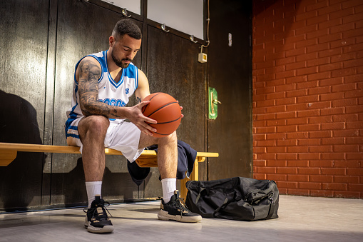 Male basketball player sitting in locker room and holding ball while waiting for sport match wide shot