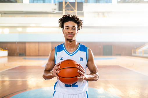 Smiling male basketball player holding ball at indoor court and looking at camera front view medium shot portrait