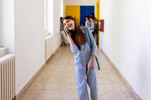 Happy Elementary Schoolgirl Talking on Smartphone in the Middle of the Corridor Near Blurred Classmates
