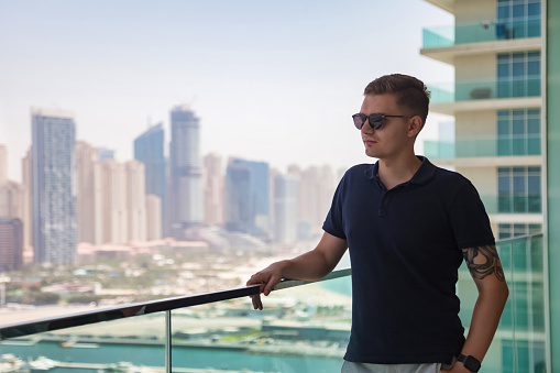 Portrait of young guy in sunglasses on skyscraper balcony with view of Dubai UAE, pensive looking away. Thoughtful male posing on terrace of tower block. Rest and leisure activity. Copy ad text space