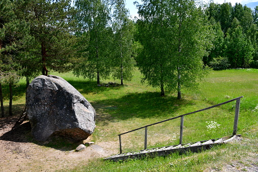 A close up on a massive rock, boulder or stone located next to a small forest or moor and a vast field, meadow, or pastureland situated near a hill with steps leading to it seen in summer