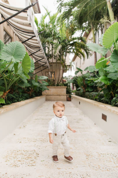 Lifestyle Candid Moment of a Cuban-American 1-Year-Old Baby Boy with Short Light Brown Hair Wearing Sage Green Bow Tie, Suspenders, White Buttondown Shirt, Pants & Brown Shoes Walking Around a Garden in Tropical Palm Beach, Florida in May of 2023 estilo de vida momento sincero de un bebé cubanoamericano de 1 año con cabello castaño claro corto con pajarita verde salvia, tirantes, camisa blanca abotonada, pantalones y zapatos marrones caminando por un jardín en tropical palm beach, florida en ma