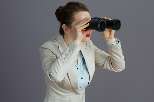 trendy female employee in a light business suit with binoculars against grey background looking into the distance.