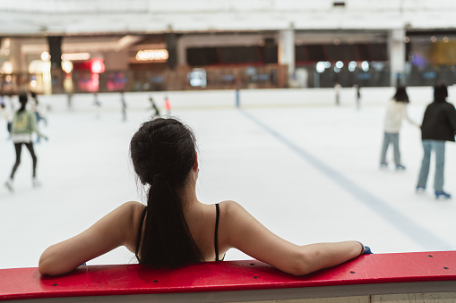 Back view of an Asian female figure skater in black leotard resting by the skating rink