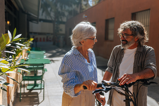 A mature married couple is outside in the city, standing next to each other with their bicycle and talking.