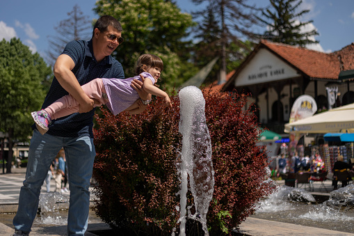 The child is playing with the water on the fountain. The man is holding his daughter, and helping her to touch the water.