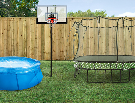 Summer outdoor fun family activities including an inflatable pool, a basketball hoop & a trampoline in a backyard with fence and green lawn