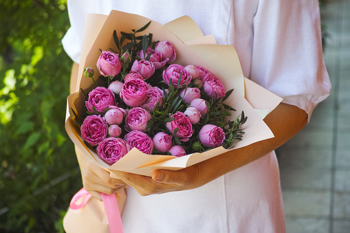 A woman wearing white dress holding a bouquet of pink peony roses in her hands outdoors.