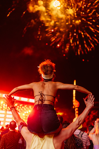 A girl is sitting on a mans shoulders at a concert in an open air venue. They are enjoying the fireworks and music.