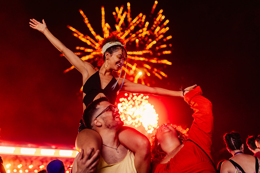 A young Caucasian girl is sitting on her boyfriends shoulder at the open air festival while he's dancing and she is holding her friends hand as she's standing next to him.