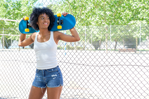 portrait of smiling young afro girl standing in front of a metal fence with a blue skateboard behind her head