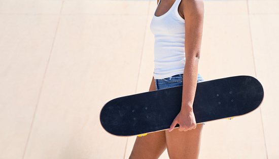 unrecognizable young afro girl in profile in white t-shirt and blue shorts in a skate park with a skateboard in her hand with copy space on the left