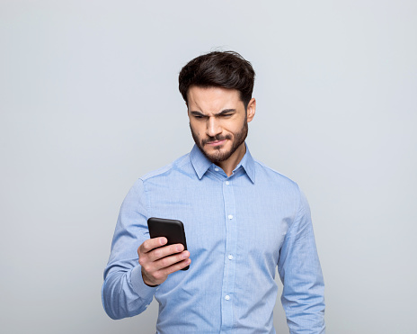 Portrait of thoughtful manager wearing blue shirt using smart phone. Studio shot, grey background.