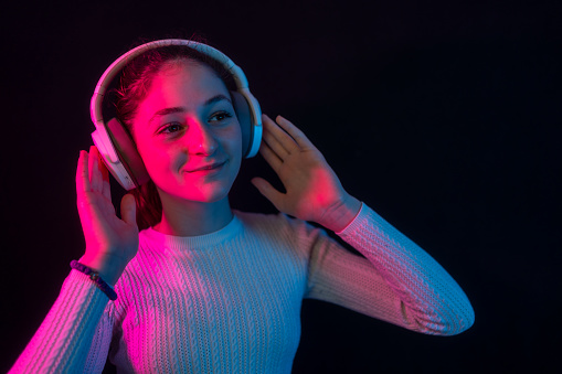 Fashion hipster girl smiles and wear headphones listening to music over color neon background at studio. enjoying listening to music in headphones, smiling, isolated on black background
