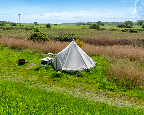 A white bell tent encamped beside the marshes near Felixstowe Ferry on the coast of Suffolk, Eastern England, on a sunny day in springtime. In the far distance, a Martello Tower can be seen behind a golf course.