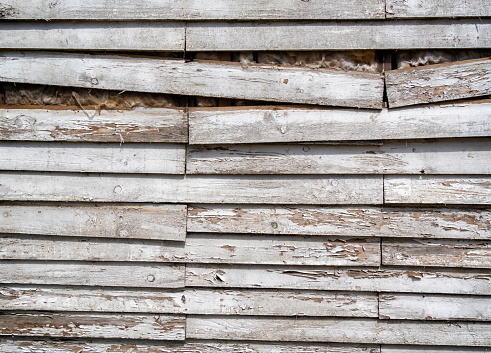 The side of an old wooden shed in sunlight, with white paint peeling away and some of the slats dropping out of place.