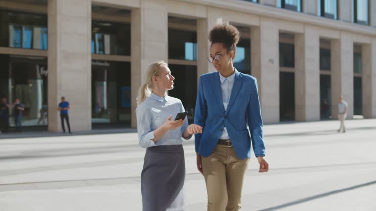 Tall and short diverse women in office clothes walking outdoors and using phone.