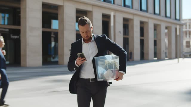 Upset businessman in suit walking outdoors with box of stuff using smartphone