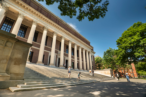 Boston, Massachusetts, USA – July 20, 2022:  Students walk by the stone steps of the grand library in Harvard Yard university campus in downtown Cambridge Massachusetts USA