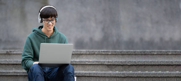 Smart young college student working on his laptop while listening to music, sitting in front of the classroom.