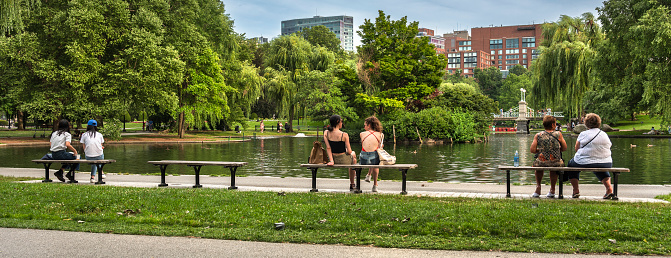 Boston, Massachusetts, USA – July 19, 2022:  Crowds of people relax on benches and picnic by the Boston Public Garden Pond in the Commons park of downtown Massachusetts USA