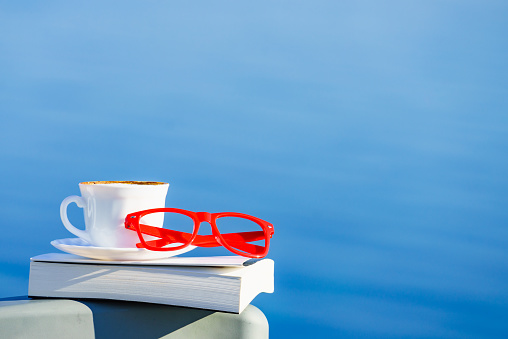 Reading on vacation. Coffee cup, book and red glasses against blue sea water. Relaxation on holidays trip. Mental health break.