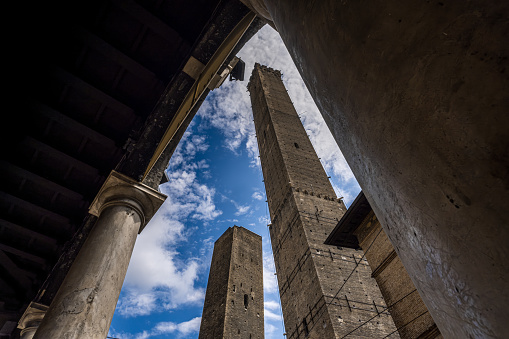 Wide-angle view of the Two Medieval Towers (Torre degli Asinelli 97 m, Torre Garisenda 48 m) in Bologna, Italy, built in the 12th century and a symbol of the city. Image taken from the arcade of Piazza della Mercanzia, view from below, on a sunny day with white clouds.