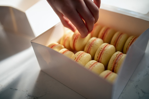 A child’s hand reaches into a box of macarons (aka French Macaroon) for a sweet treat.  A meringue based delicacy.