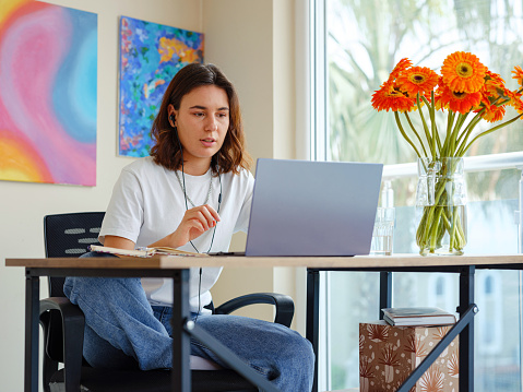 Beautiful Latin young woman in casual clothing using laptop and smiling while working indoors. lady work on computer listening music with earphones.