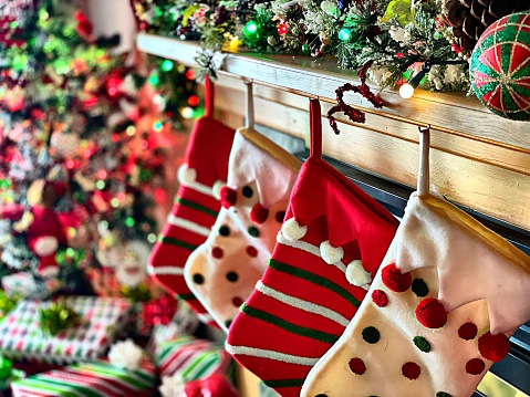 Full frame Christmas holiday background. Presents under decorated tree blurred in corner. Selective focus on foreground stockings hanging from mantle. Bright colors red and green. Home interior decorated for Christmas.