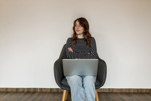 Copy space shot of charming young brunette sitting in armchair against a white background, arms crossed, using laptop. She is looking away, smiling and contemplating.