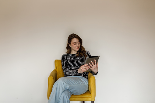 Portrait of charming young woman sitting in a yellow armchair against white background, legs crossed, relaxing and scrolling via digital tablet that she is using.