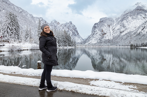 Woman in black clothes posing and smiling by a beautiful view of Toblacher See (Dobbiaco Lake) in a snowy winter day. Reflections of mountains and trees on water; Dolomites, Italy