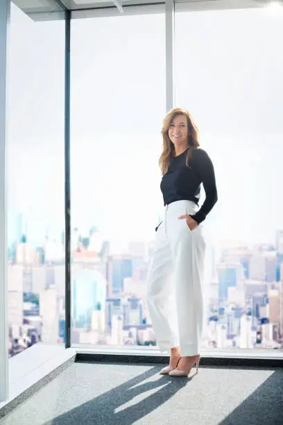 Full length shot of an attractive businesswoman wearing sweater and trousers while standing at window and looking at camera.