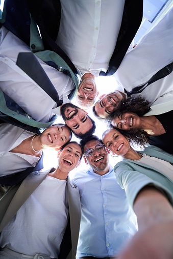 Multiethnic group of work colleagues embraced in circle, face down taking a selfie smiling and looking at the camera. Vertical image, POV business people doing team building.