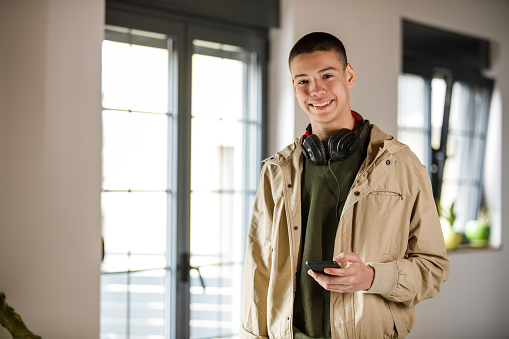 Copy space shot of charming teenage boy, headphones around his neck, standing in a room, holding smart phone, smiling at camera with confidence.