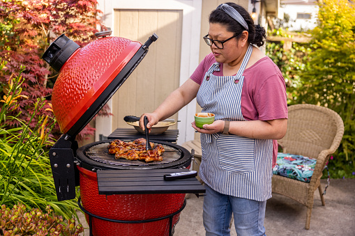 High quality stock photo of an Asian-American woman preparing and barbecuing chicken for a summer time party at home in the backyard.