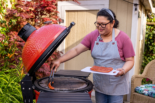 High quality stock photo of an Asian-American woman preparing and barbecuing chicken for a summer time party at home in the backyard.