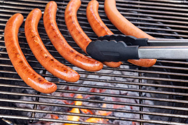 A Bratwurst or hot dog is being displayed on tongs cooked on a Flaming grill, with smoke and flames rising from the grill. A common outdoor activity during the summer months