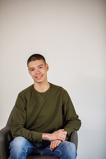 Portrait of charming teenage boy sitting in a chair against a white background, looking at camera and smiling with confidence.