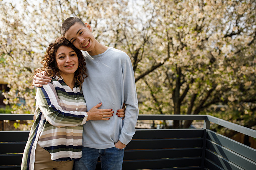 Copy space shot of caring teenage boy and his mid adult mother standing on the sunny balcony, embraced, having bonding moments. They are looking at camera and smiling happily.