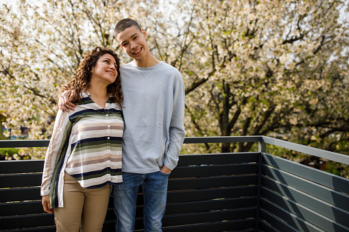 Copy space shot of caring teenage boy and his mid adult mother standing on the sunny balcony, embraced, having bonding moments. Boy is looking at camera and smiling while mother is looking at him.