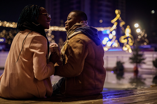 Rear view of caring young woman surprising her loving boyfriend with a Christmas gift bag while sitting at the Christmas park in the evening, enjoying the view of the glistening Christmas lights.