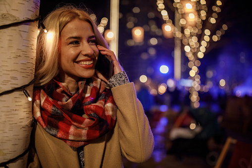 Portrait of joyful young woman standing by the pole decorated with Christmas lights, at the Christmas park, talking on the phone while waiting to meet a friend. She is looking away, smiling and contemplating.